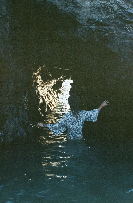 A woman wading through dark water toward the light at the mouth of a sea cave.